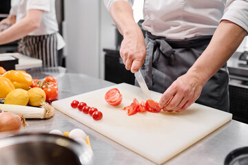 Close-up of young cook in apron using knife to cut fresh tomatoes on cutting board at table