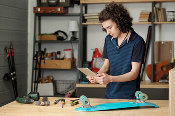 Contemporary guy in casual clothes looking through online adverts while standing by table with blue skateboard and handtools in garage