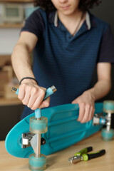 Hands of teenage guy with wrench or screwdriver fixing wheels of blue skateboard or repairing it while working by table or workbench