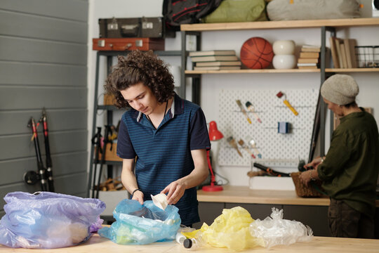 Youthful guy in casualwear putting plastic food containers into one of several cellophane sacks while sorting waste by table in garage