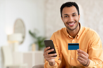 Smiling young guy using smartphone and credit card at home