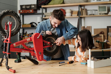 Young man holding hand of his little daughter while checking wheels of her bicycle or repairing it...