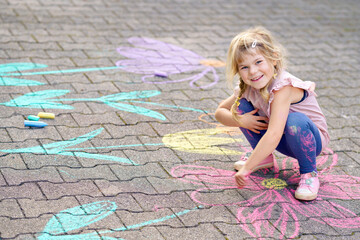 Little preschool girl painting with colorful chalks flowers on ground on backyard. Positive happy toddler child drawing and creating pictures on asphalt. Creative outdoors children activity in summer.