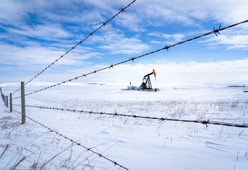 An oil pump jack working in a snow covered agriculture field along a barbed wire fence on the...