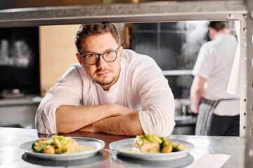 Portrait of young professional cook in eyeglasses looking at camera behind the counter with ready dishes