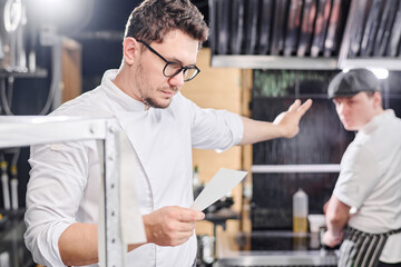 Young serious chef in eyeglasses reading order and gesturing his assistant to prepare dish for two in kitchen