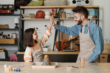 Happy little girl and her father in aprons and protective eyeglasses making high-five by table with...