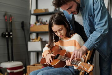 Little girl in eyeglasses learning to play guitar while young man bending over her and keeping...