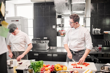 Young chef cutting tomatoes on cutting board and discussing menu with his assistant during their...