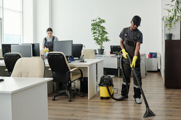 Young African American man in workwear cleaning floor of contemporary office with vacuum cleaner...