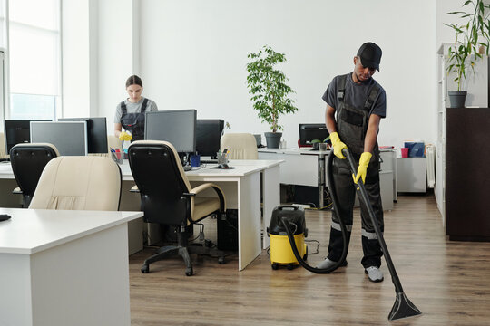 Young African American man in workwear cleaning floor of contemporary office with vacuum cleaner while standing against workplaces