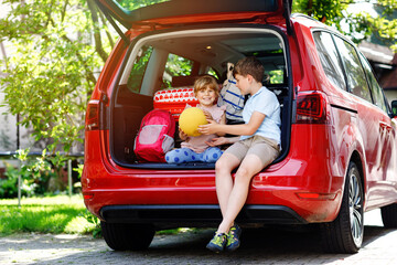 Two children, school boy and preschool girl sitting in car trunk before leaving for summer vacation with parents. Happy kids, siblings, brother and sister with suitcases and toys going on journey
