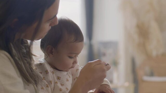 Happy beautiful woman sitting at kitchen table and feeding her baby daughter with apple puree. Young mother holding infant on lap while child eating fruit mash. Parenthood, motherhood concept