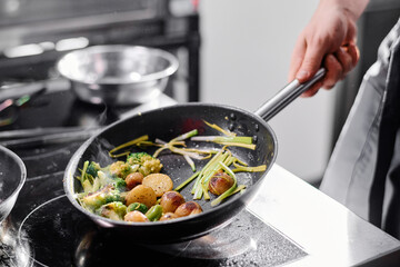 Close-up of professional cook holding pan over the cooker and frying potatoes with vegetables