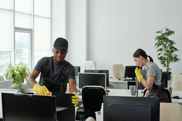 Young male staff of modern cleaning service company wiping computer monitor while his female...