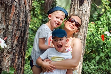 Mother and little boys stand leaning on pine trunks in hotel park. Woman in sunglasses holds toddler and preschooler in hats on sunny day closeup