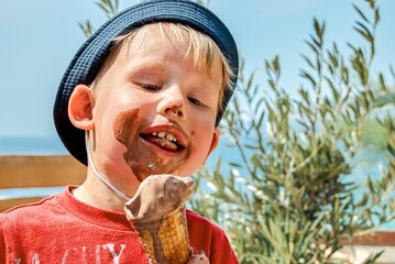 Blond boy in panama hat enjoys eating chocolate ice cream getting dirty. Cute toddler sits on bench...