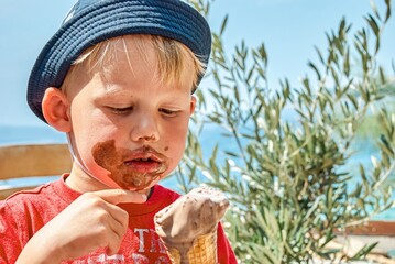 Blond boy in panama hat enjoys eating chocolate ice cream getting dirty. Cute toddler sits on bench against sea on vacation in Omis closeup - 500970898