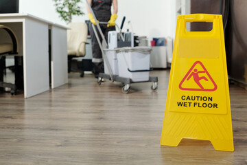 Caution of wet floor on yellow signboard in openspace office against female cleaner pushing janitor cart with buckets and mop