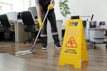 Yellow plastic signboard with caution warning about wet floor and female cleaner in black uniform...