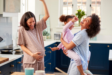 Pregnant Family With Two Mums Wearing Pyjamas Dancing In Kitchen With Daughter
