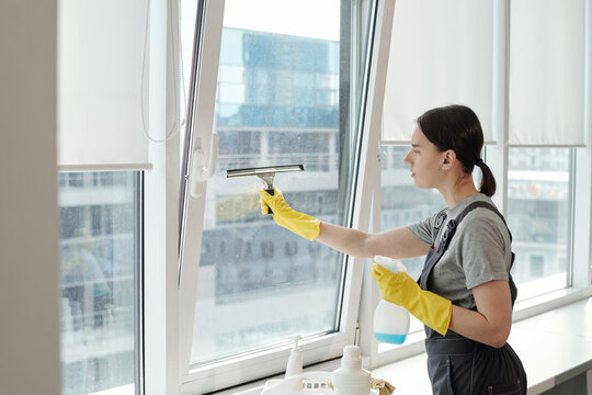 Side view of young female cleaner in workwear rubbing large office window with silicone spatula while carrying out her work