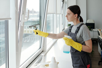 Young female staff of cleaning company washing large office window with special silicone spatula...