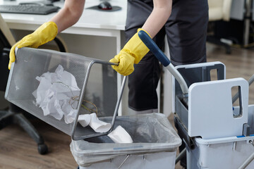 Gloved hands of female cleaner throwing trash from garbage bin into plastic bucket on janitor...