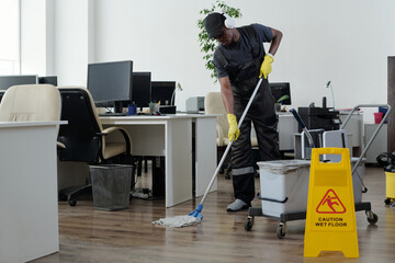 Contemporary young black man in workwear cleaning floor in openspace office in front of yellow...