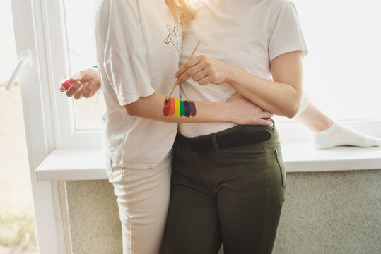 Lesbian couple hugs and painting together rainbow and holds brush. No face. Romantic embrace
