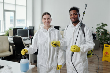 Two happy young intercultural workers of cleaning service in protective overalls and gloves...