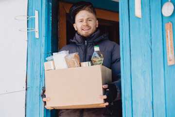 Young caucasian refugee man is happy with box of humanitarian aid, meets on threshold of house...