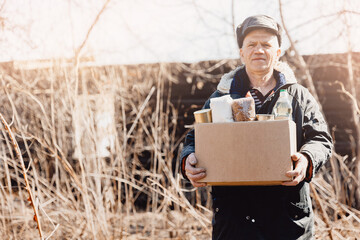 Old man senior is holding cardboard box of donations canned food, looking camera. Concept humanitarian aid to refugees