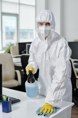 Female worker of cleaning service in protective workwear disinfecting desks and other surfaces in modern openspace office