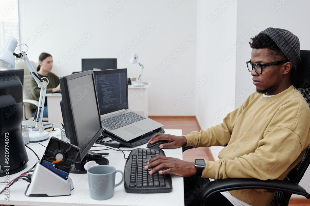 Poster Young African American it developer in casualwear decoding data while sitting in front of computer screen in modern openspace office