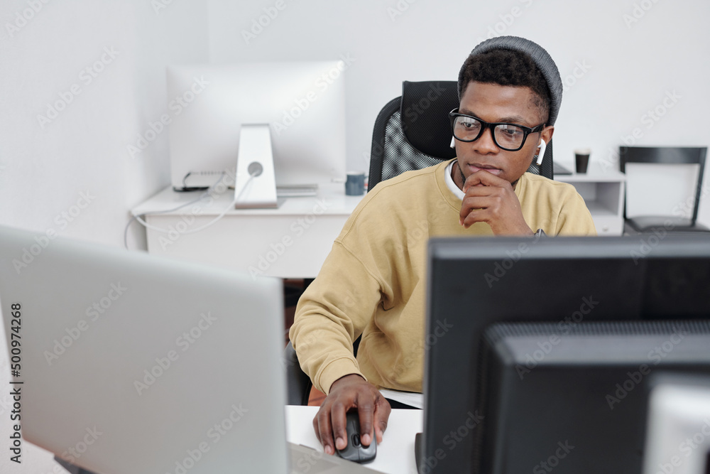 Poster young serious african american man in casualwear and eyeglasses decoding data in front of computer m