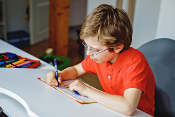 Naklejka premium Portrait of little kid boy with glasses at home making homework, writing and learning. Little child doing exercise, indoors. Elementary school and education, home schooling concept.