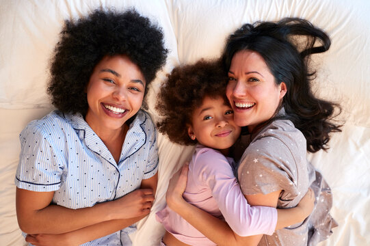 Overhead Portrait Of Family With Two Mums Wearing Pyjamas Playing On Bed At Home With Daughter