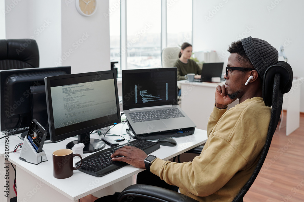 Poster Side view of young serious lack man concentrating on decoding data while sitting in front of computer monitor by workplace in office