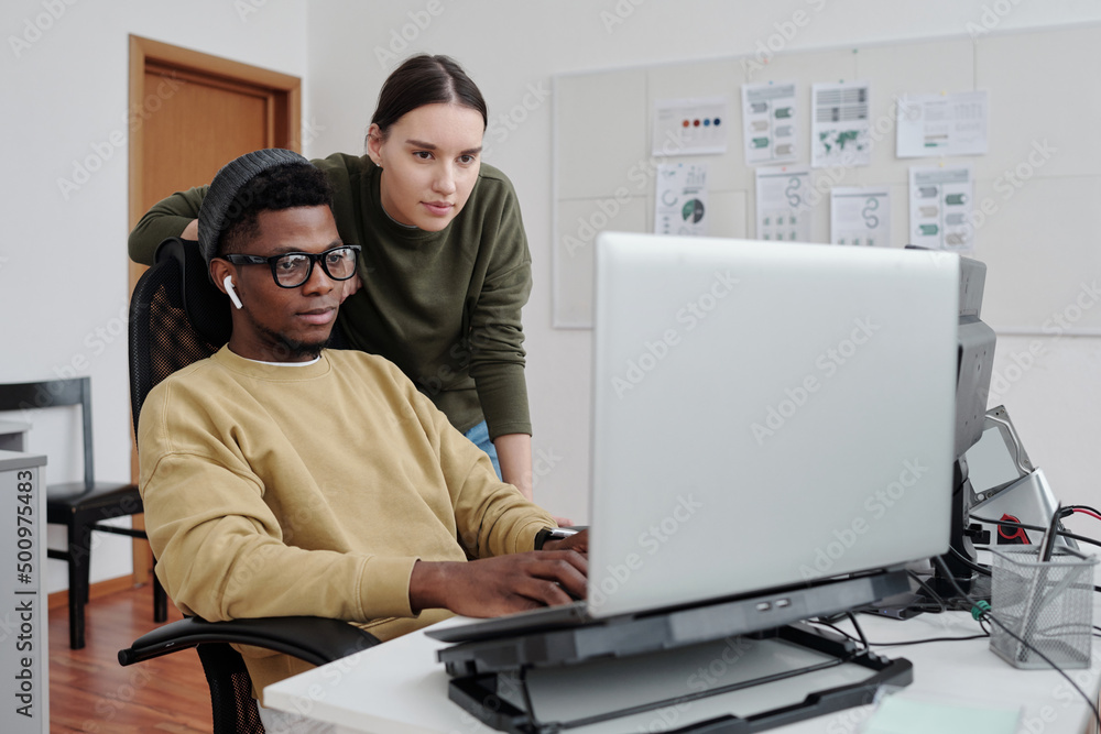Poster two contemporary programmers decoding data on laptop screen by workplace while young african america