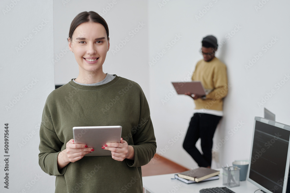 Poster young successful female programmer or manager with tablet looking at camera while standing against a