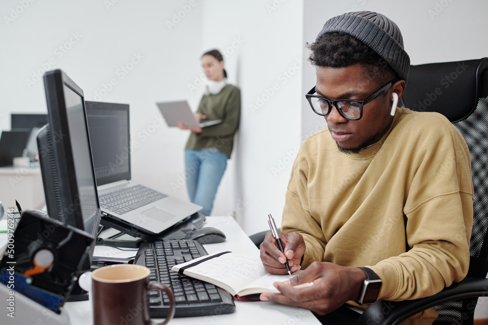 Aufkleber serious african american guy in eyeglasses and casualwear making notes in copybook while sitting by 