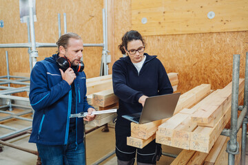 Two carpenters are standing in the workshop looking at a laptop. They are consulting with each...