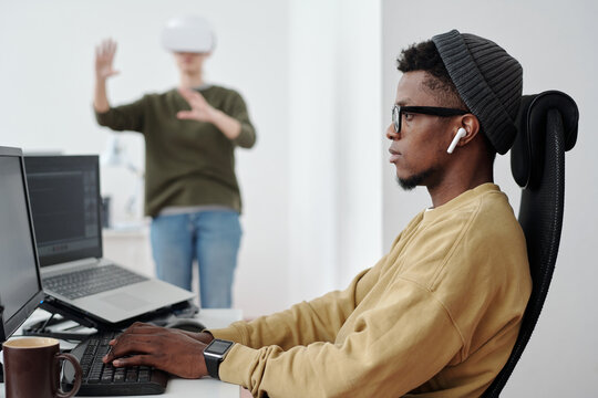 Side view of young serious businessman in earphones, eyeglasses and casualwear decoding data while looking at computer screen