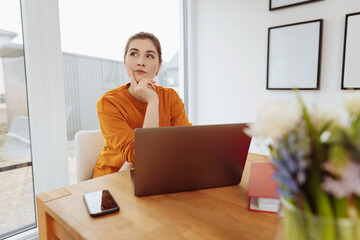 young thoughtful woman sits at wooden table in front of laptop in living room