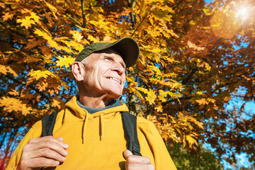 Elder hiker with smile and with backpack standing against oak tree leaves at autumn forest. Active and healthy life style concept.