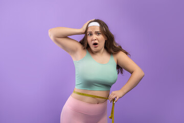 Obraz na płótnie Canvas Surprised sad young european plus size woman in sportswear with open mouth measures waist with measuring tape