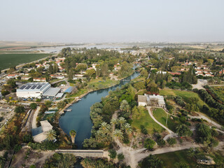 Aerial panorama view of Kibbutz Nir David in Northern Israel with nahal Amal river turquoise water between houses and palm trees surrounding agricultural land.