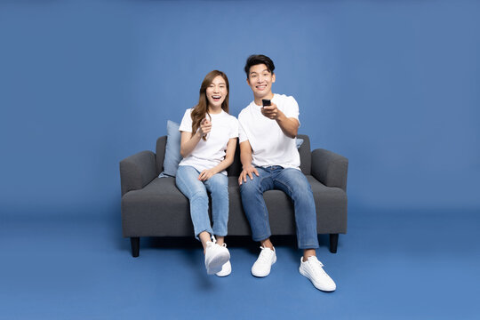 Young Asian Couple Holding Remote Tv And Watching Television On A Sofa Isolated On Blue Background