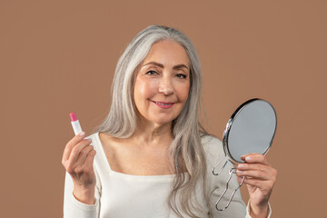 Pretty mature lady looking in mirror and applying lipstick on brown studio background. Beauty and...